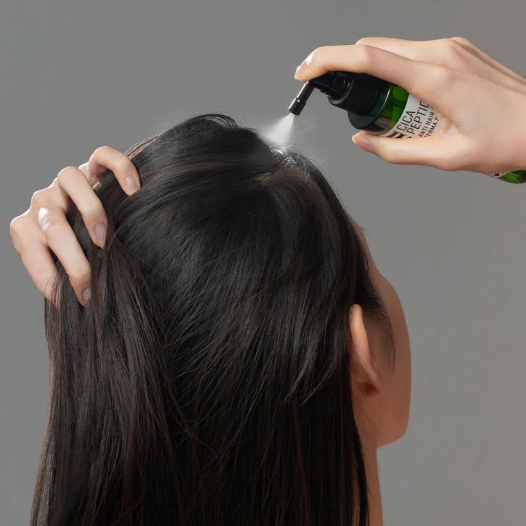 Person spraying Cica Peptide Anti-Hair Loss Derma Scalp Tonic on dark hair scalp against gray background