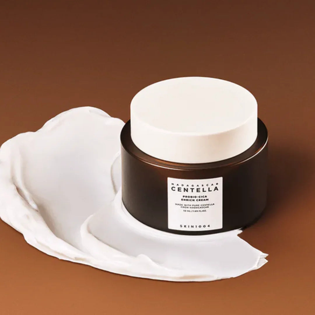 Skin1004 Madagascar Centella Probiotics Cica Enrich Cream in dark jar with white lid on brown background with cream texture